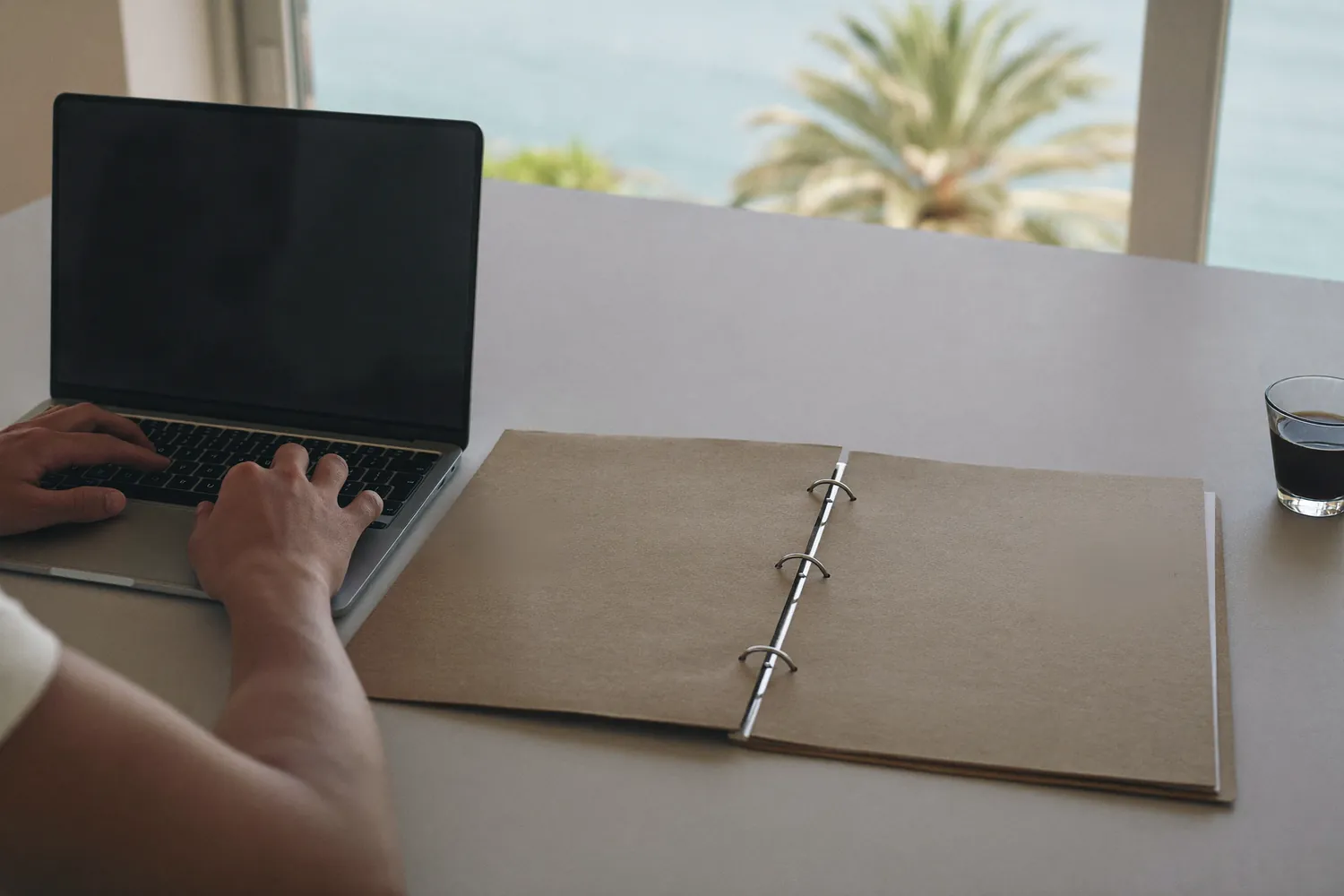 Hands at a laptop beside an open folder, sea beyond the window